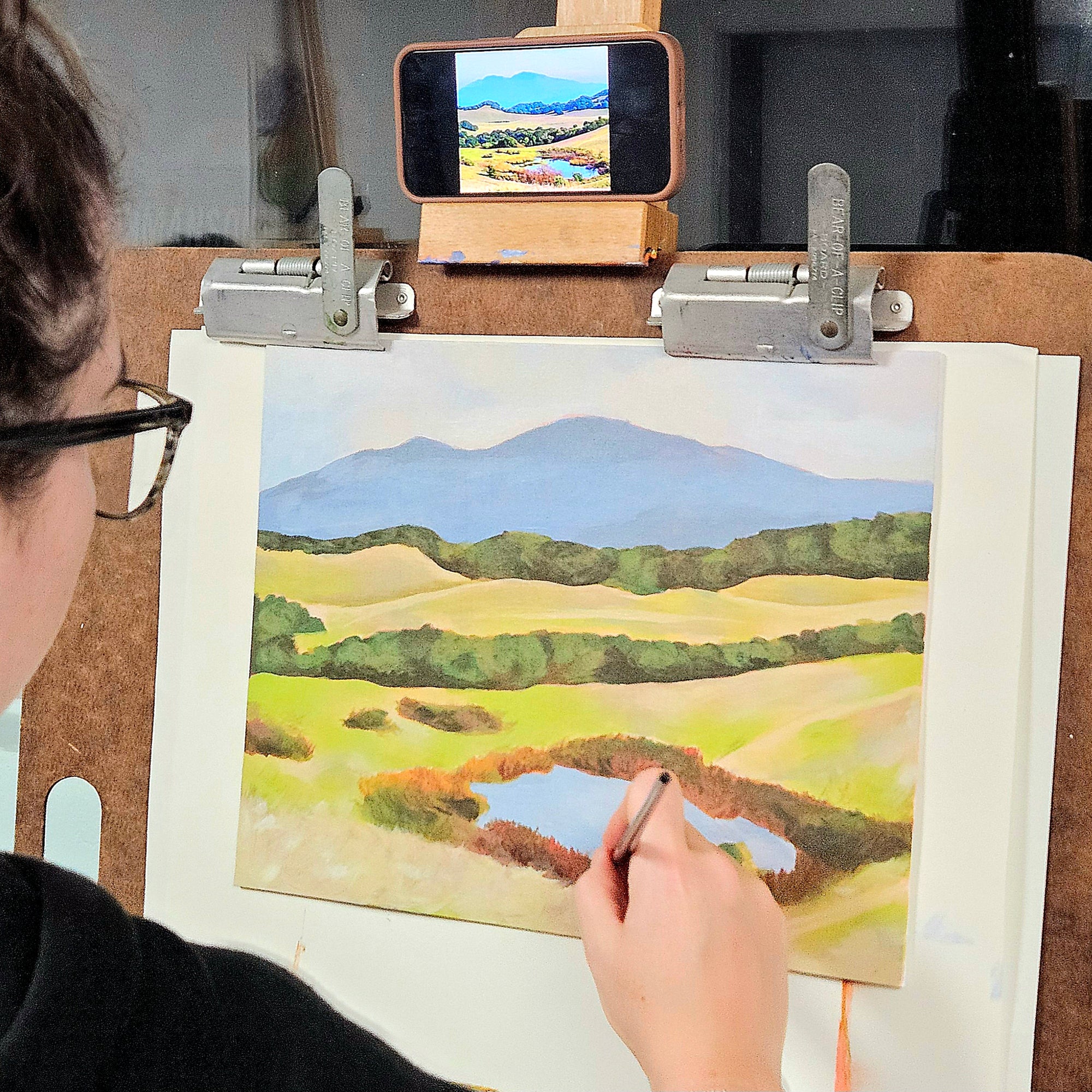 A person painting a landscape scene on a canvas mounted on an easel, with a smartphone held above the canvas showing the painted scene.