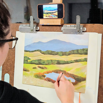 A person painting a landscape scene on a canvas mounted on an easel, with a smartphone held above the canvas showing the painted scene.