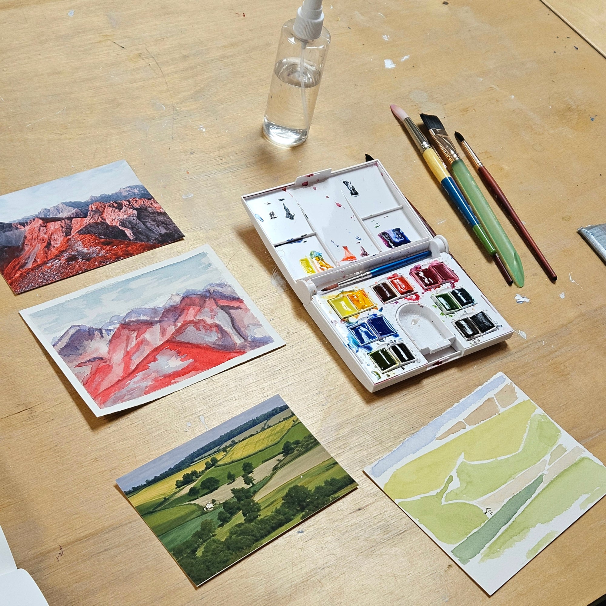 An open watercolor paint set with multiple color wells, several paintbrushes, a small spray bottle, and several painted landscape and abstract paper sketches arranged on a light wood tabletop.