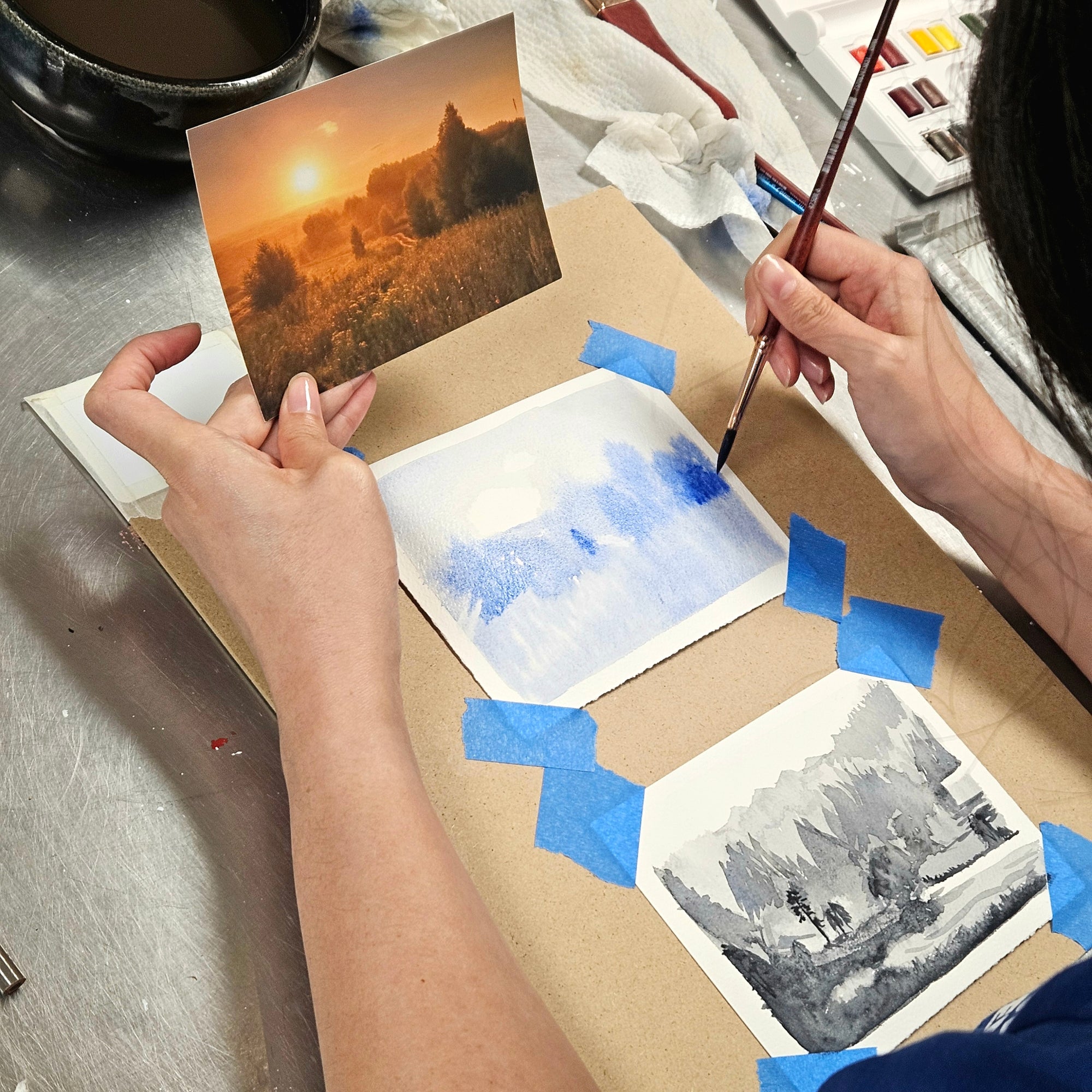A person's hands are shown painting landscape art prints on a cardboard sheet, holding a reference photo of a sunset field and using a paintbrush on blue watercolor swatches.