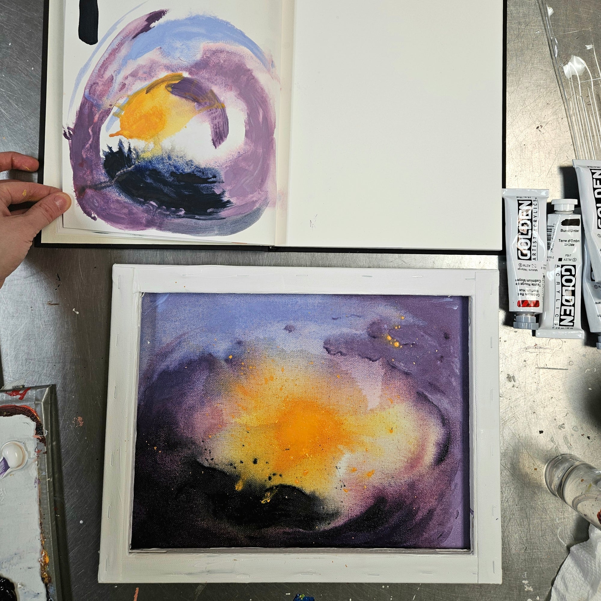 A watercolor painting in progress on a white paper sheet, showing a purple and blue spiral with a yellow bird, alongside a finished abstract painting in a white frame. Nechrol watercolor tubes and paint brushes are visible on a metal workbench.