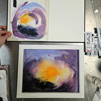 A watercolor painting in progress on a white paper sheet, showing a purple and blue spiral with a yellow bird, alongside a finished abstract painting in a white frame. Nechrol watercolor tubes and paint brushes are visible on a metal workbench.