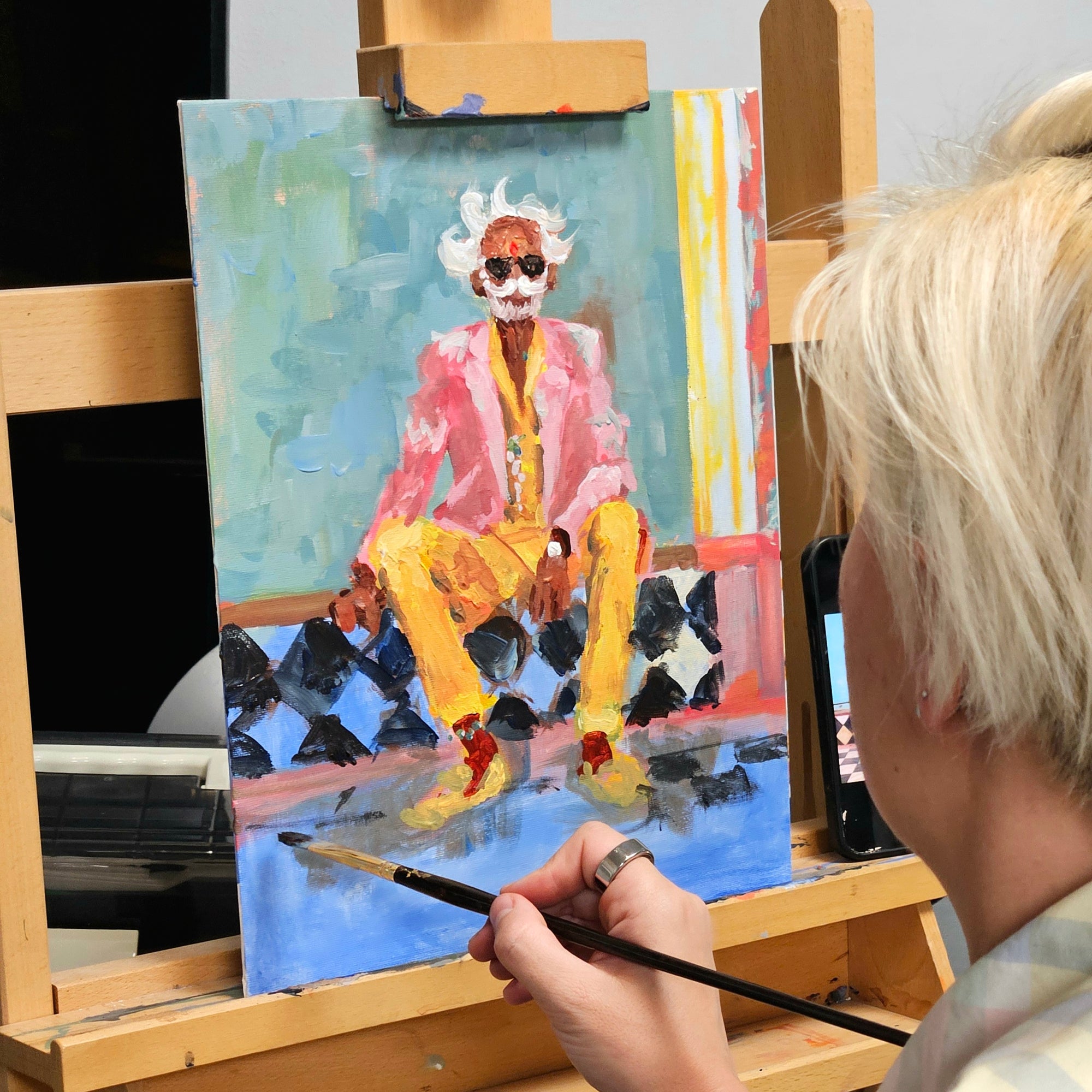 A person painting a portrait of Albert Einstein on a canvas set on an easel, with the artist's hand and blonde hair visible in the foreground.