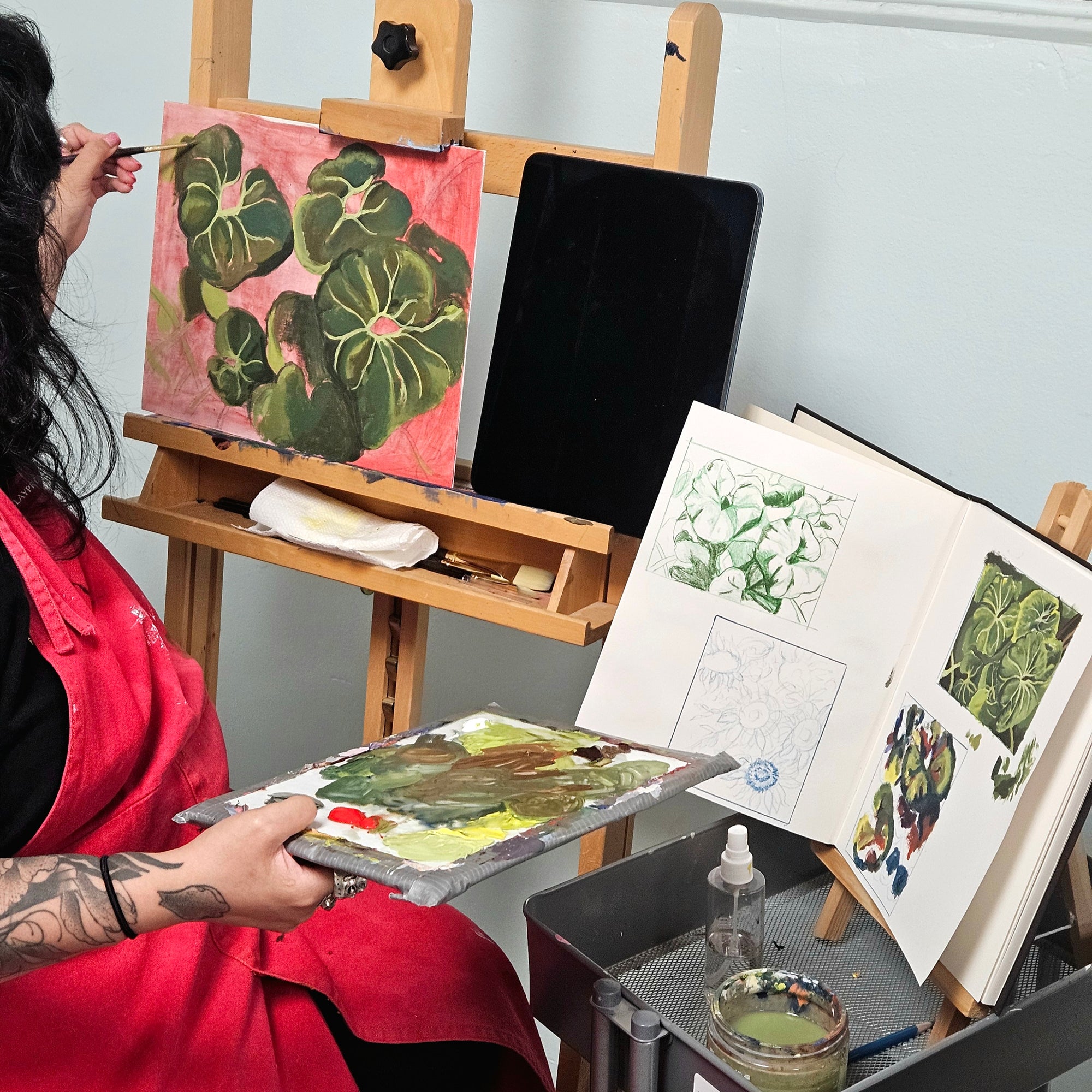 A person in a red painter's apron sits at an easel, holding a paint palette with green botanical artwork. A finished painting of green leaves on pink paper is displayed on the easel, alongside an open sketchbook and a tablet, in a studio setting.