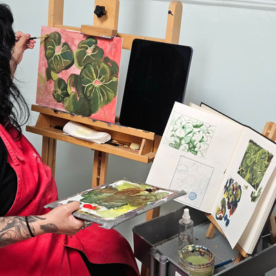 A person in a red painter's apron sits at an easel, holding a paint palette with green botanical artwork. A finished painting of green leaves on pink paper is displayed on the easel, alongside an open sketchbook and a tablet, in a studio setting.