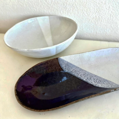 Two handmade ceramic dishes on a light cream surface against a textured off-white wall: a shallow white bowl in the background and a larger speckled white plate with a glossy dark purple and brown glazed foreground piece.