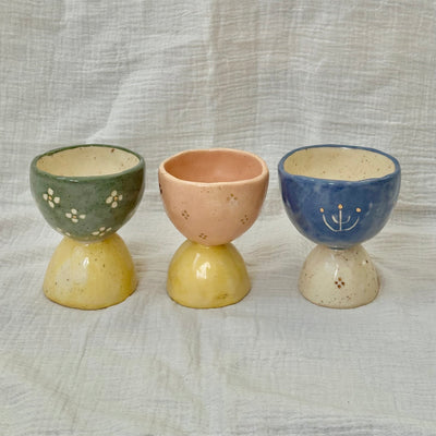 Three ceramic egg cups in green, pink, and blue on a textured fabric background